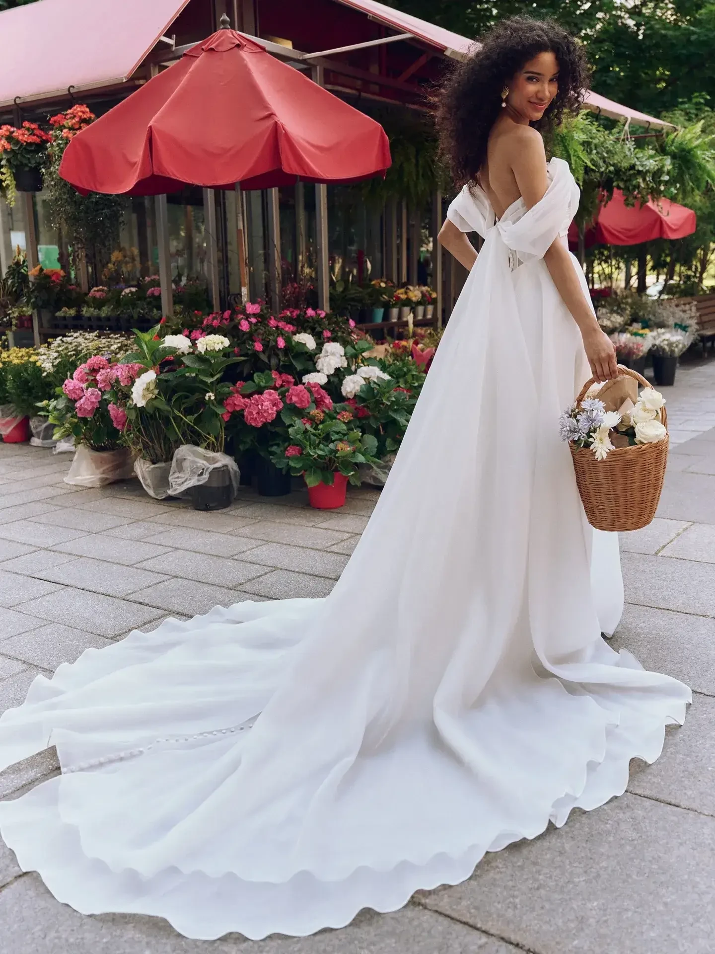 A woman in a flowing white gown gracefully walks along a sidewalk, holding a wicker basket. She smiles against a backdrop of vibrant flowers and red canopies.
