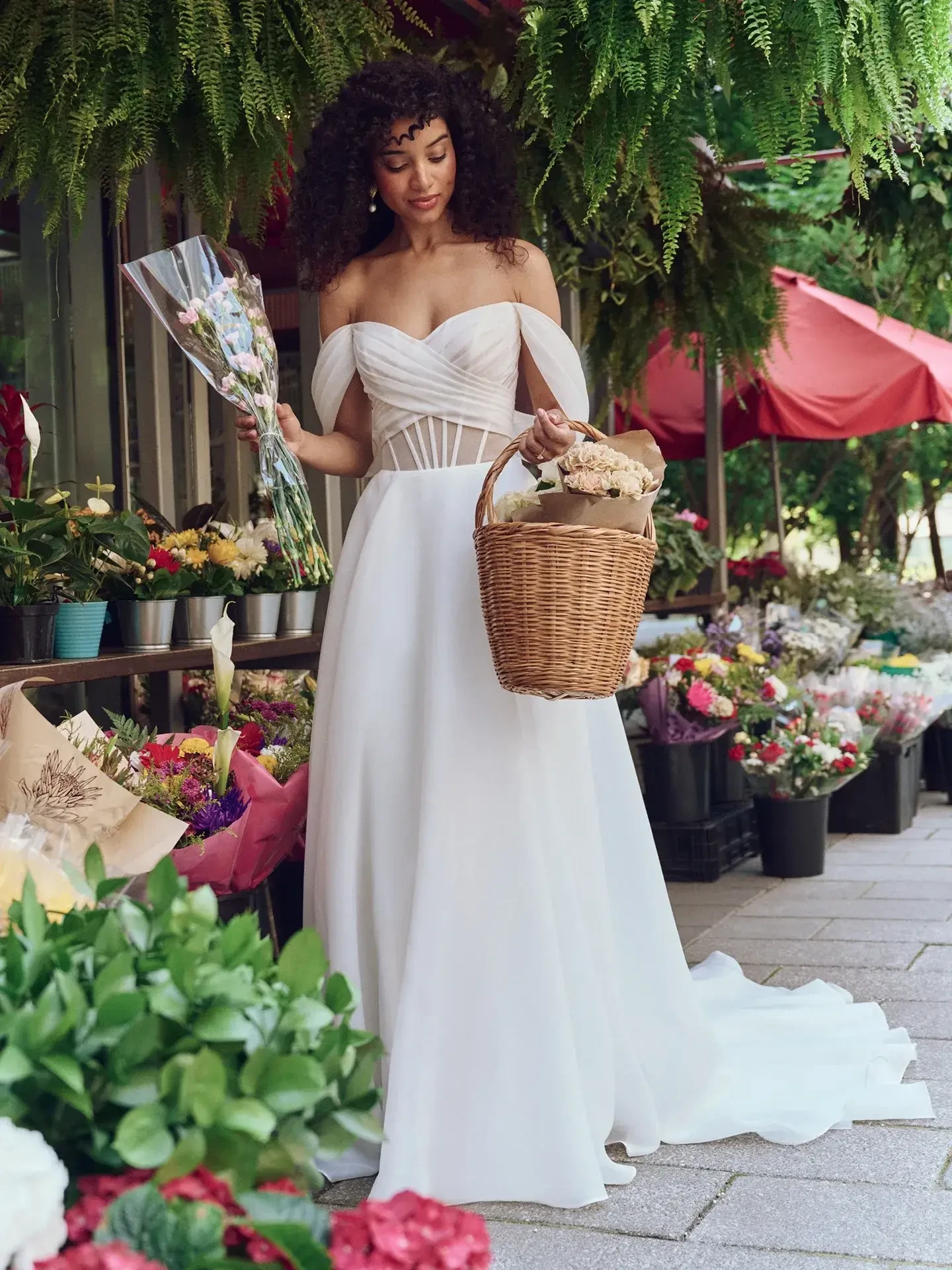 A woman in an elegant, off-the-shoulder white dress holds a wicker basket of flowers in a vibrant outdoor flower market, exuding a serene and romantic vibe.