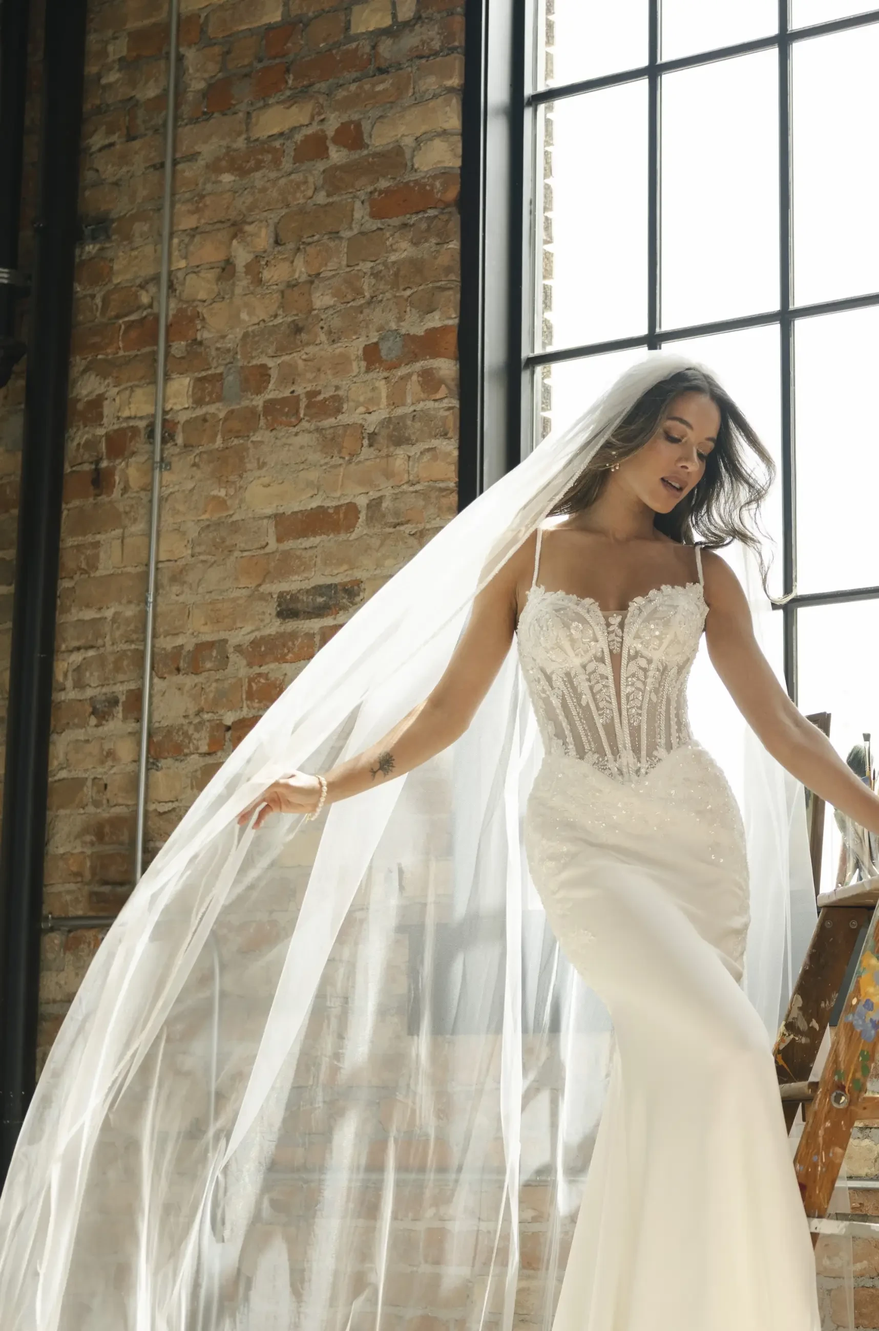 A woman in a flowing white wedding dress with intricate lace details and a long veil, stands gracefully near a sunlit brick wall, exuding elegance and joy.
