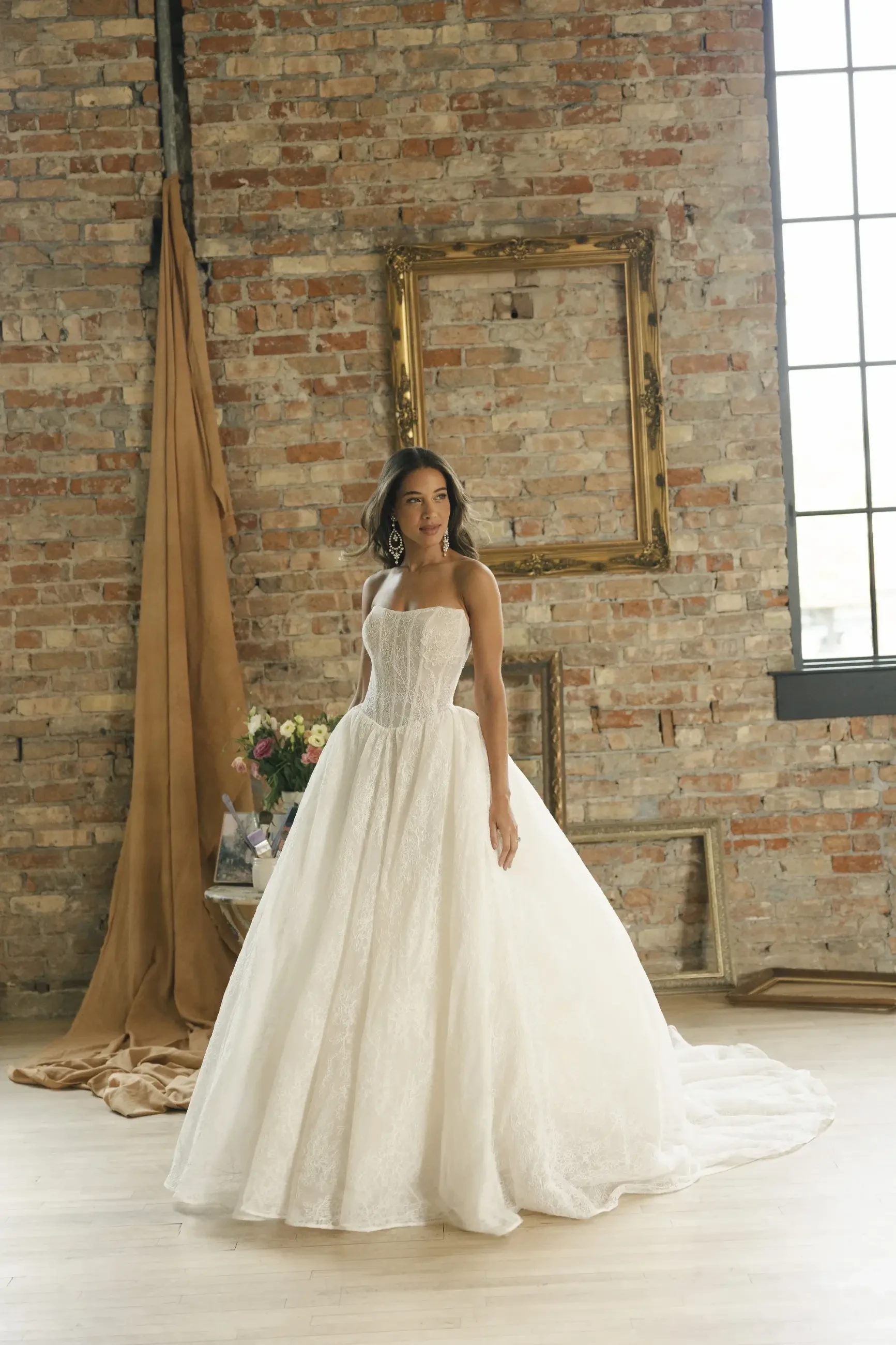 A woman in a white, strapless wedding gown stands in a rustic room with brick walls and tall windows. Frames and draped fabric add elegance.
