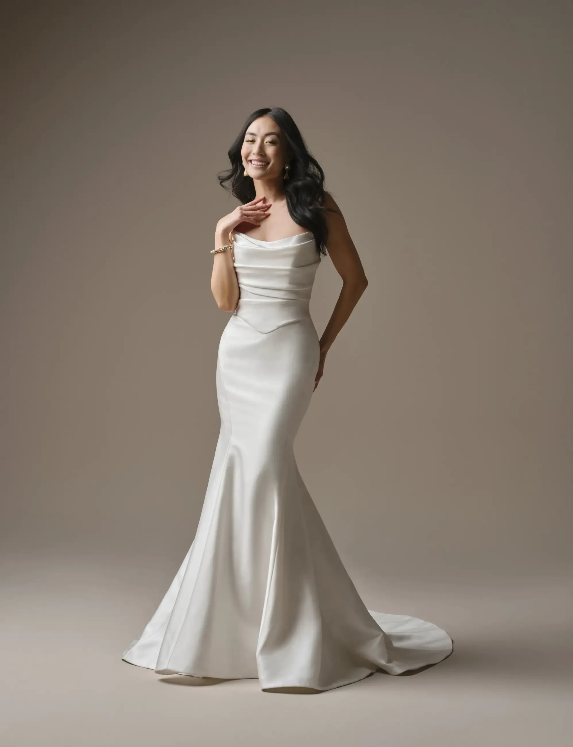 Elegant woman in a sleek, white mermaid gown, poses with a joyful smile. Her dark wavy hair cascades down, and a soft, neutral background adds warmth.