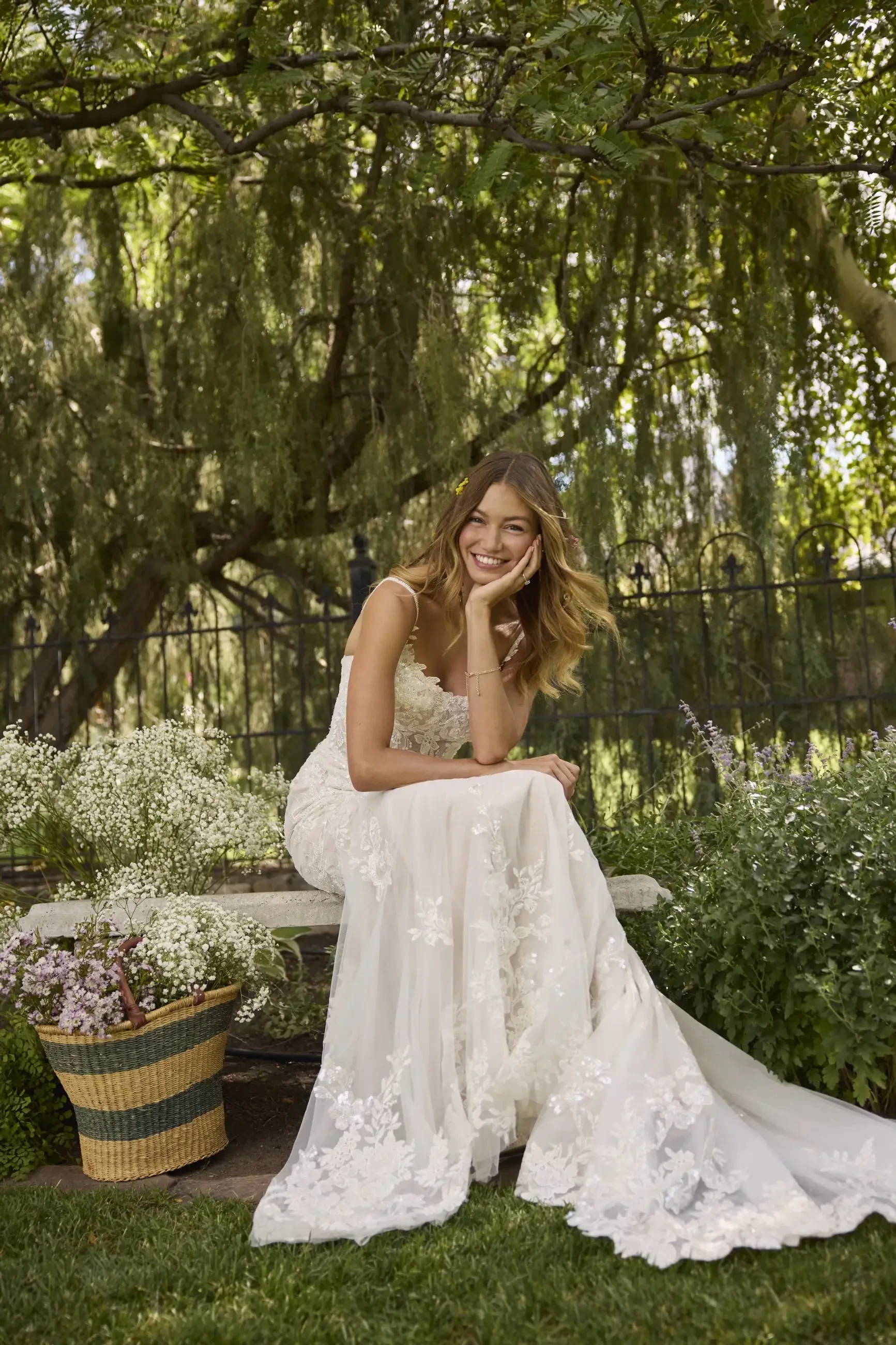 A woman in a lace wedding dress sits on a garden bench, smiling amid lush greenery and flowers. A woven basket nearby adds a rustic touch.