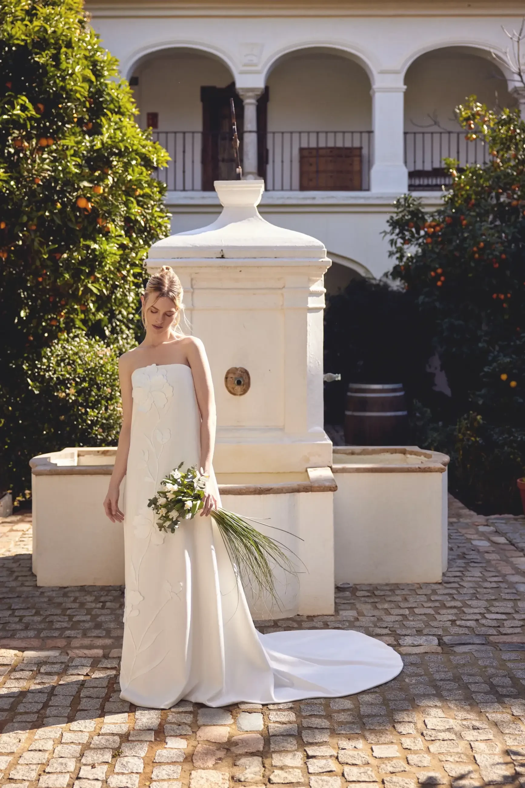 A bride in a simple, elegant white strapless wedding gown stands near a decorative fountain in an outdoor setting, holding a bouquet of flowers. The backdrop features greenery and orange trees.