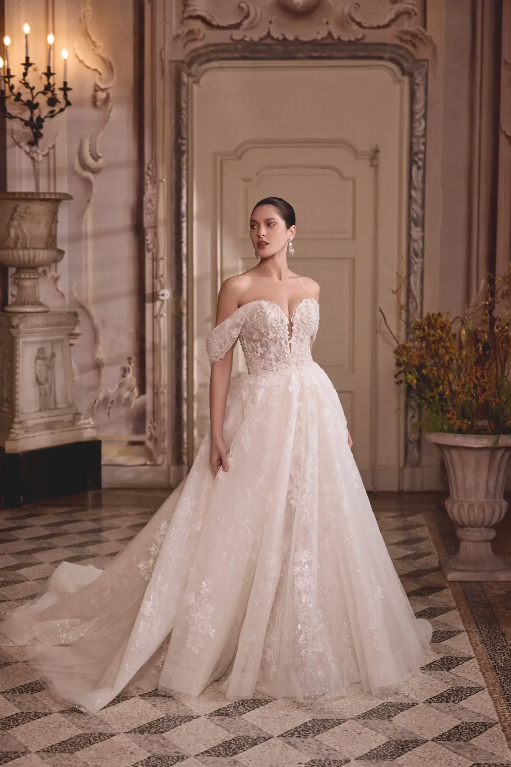 A woman in an elegant off-the-shoulder wedding dress with lace and floral embellishments, standing in a lavish indoor setting with ornate decor and tile flooring.