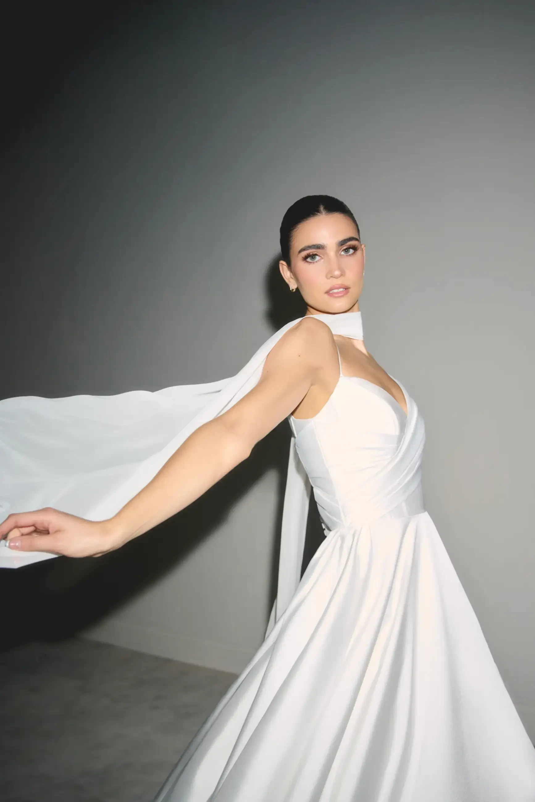 A person wearing a white dress with a flowing cape, posing with an elegant gesture. The background is simple and minimalistic.