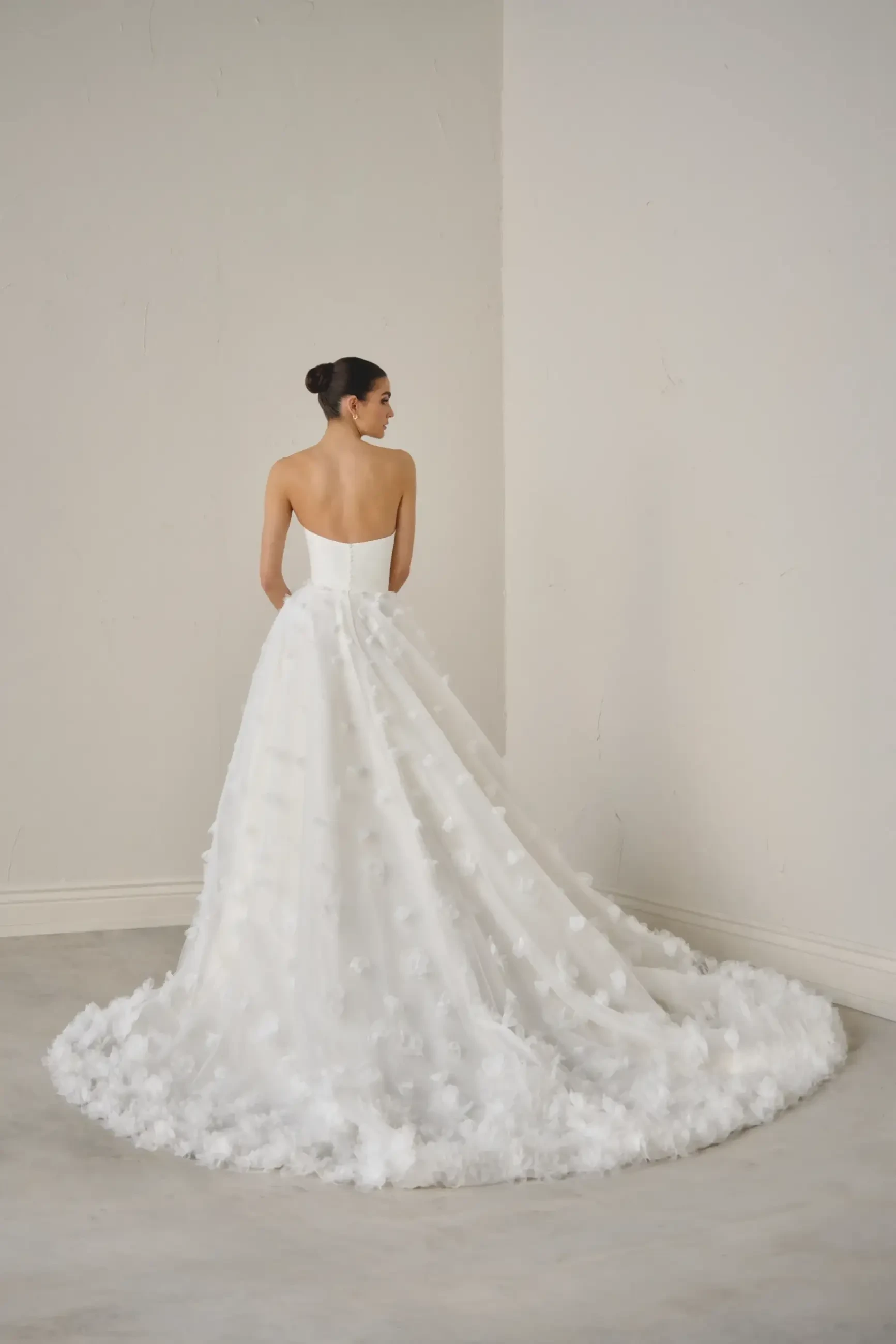 A woman in a strapless, white ball gown with a long, floral-embroidered train is standing with her back to the camera in a minimalist room. Elegant and serene atmosphere.