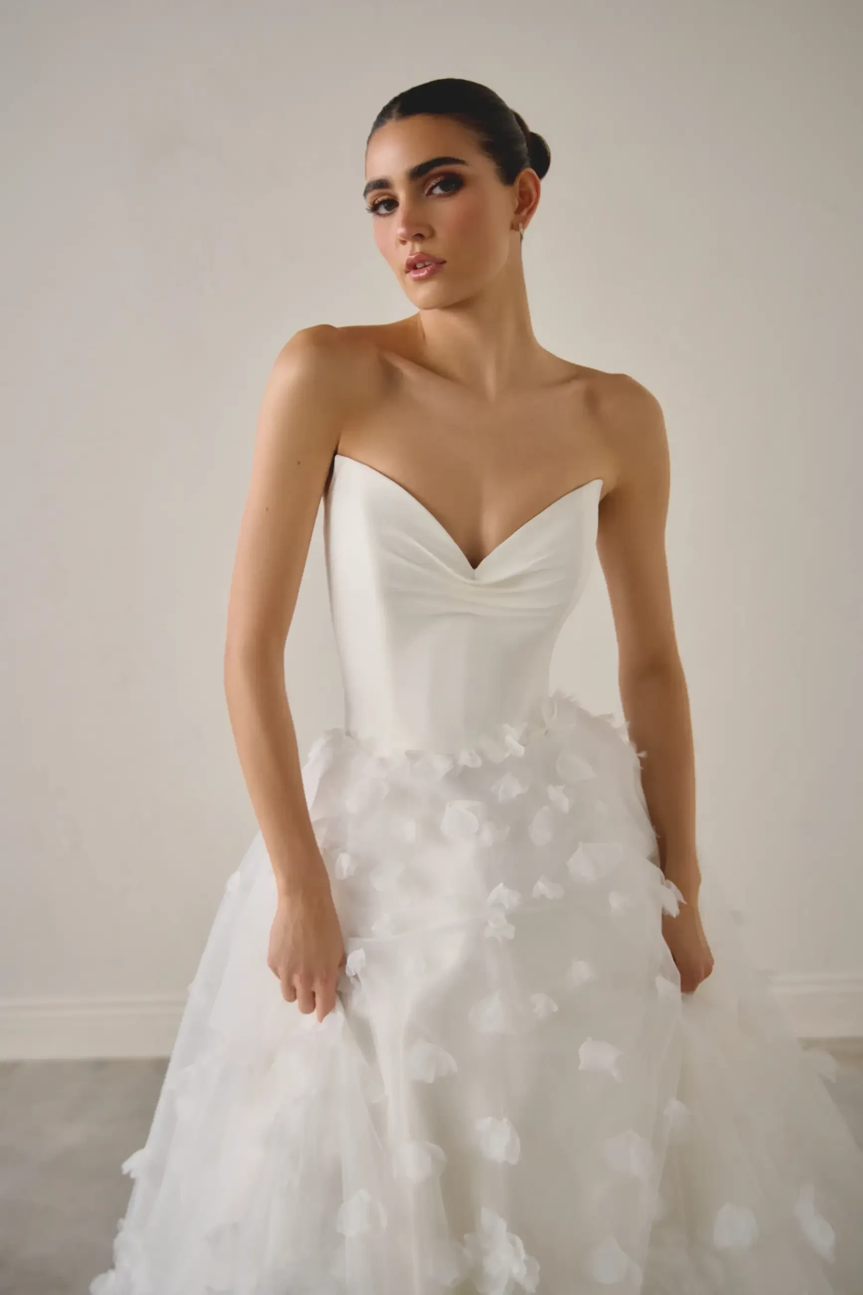 Woman in an elegant, strapless white wedding gown with floral details, standing against a plain background, conveys elegance and sophistication.