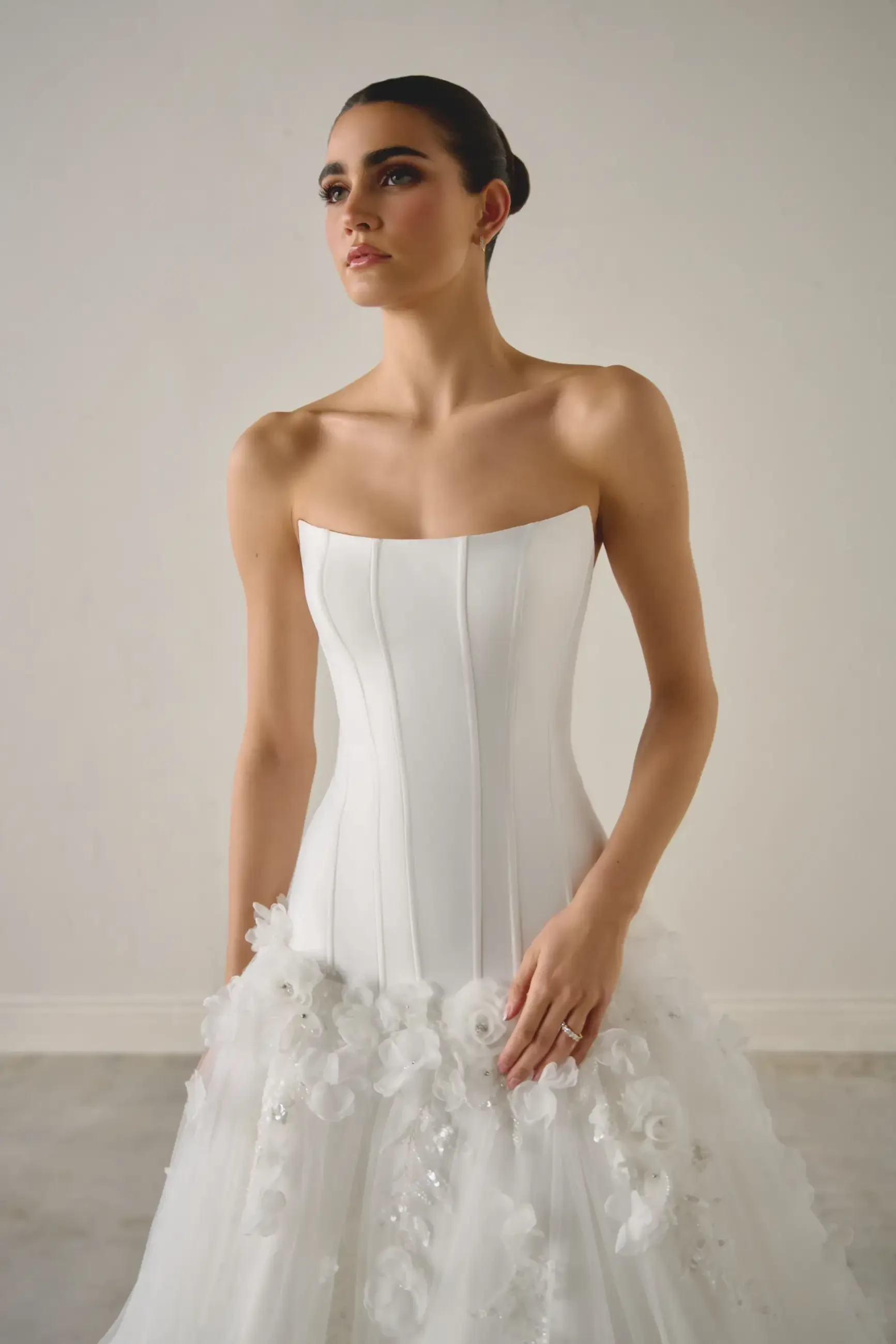 A model wearing a strapless white wedding dress with intricate designs at the hem, standing in a neutral-colored setting.