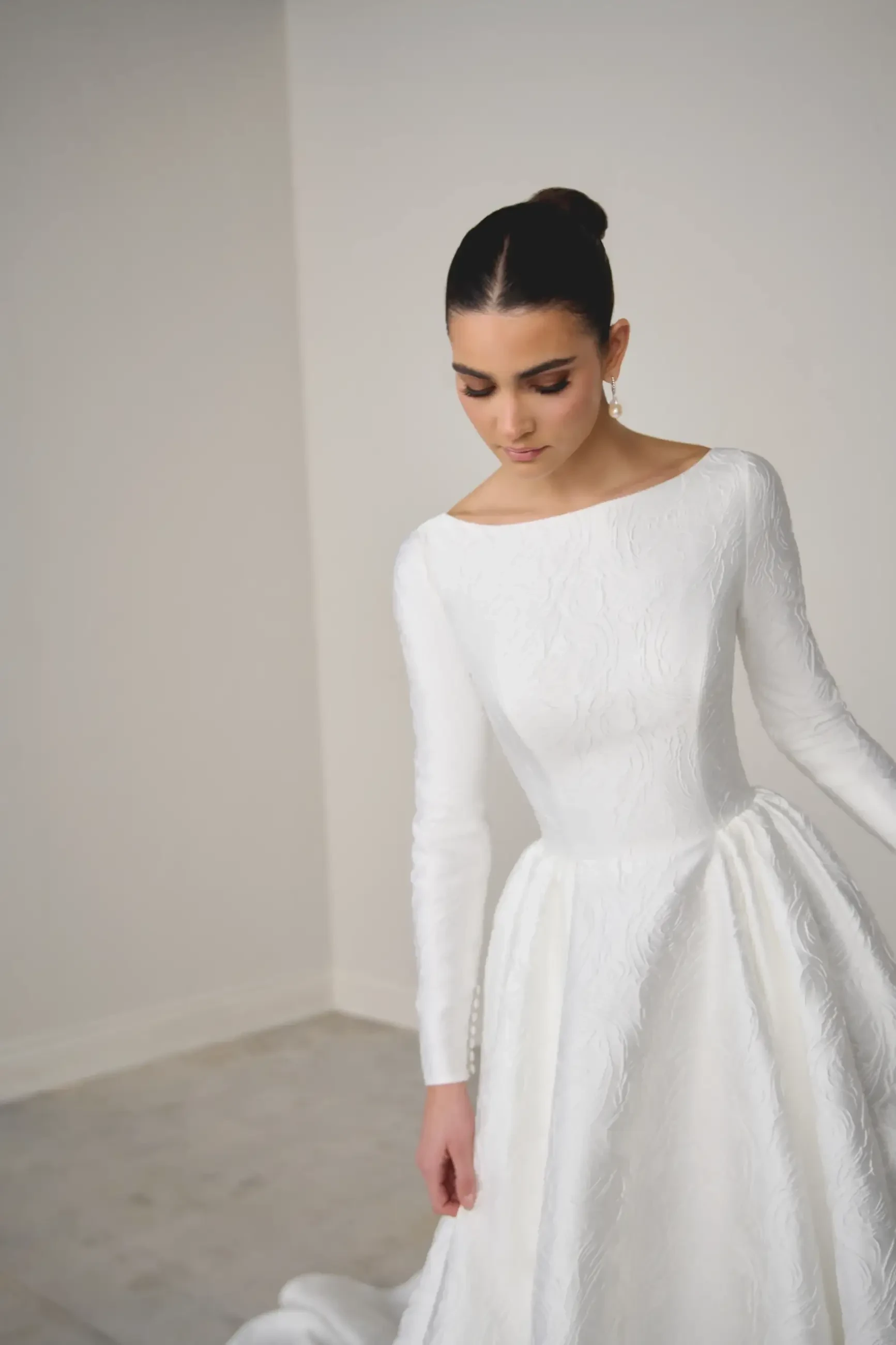 A woman wearing a long-sleeved, white wedding dress, standing and looking down. The dress has a structured bodice and a flowing skirt.