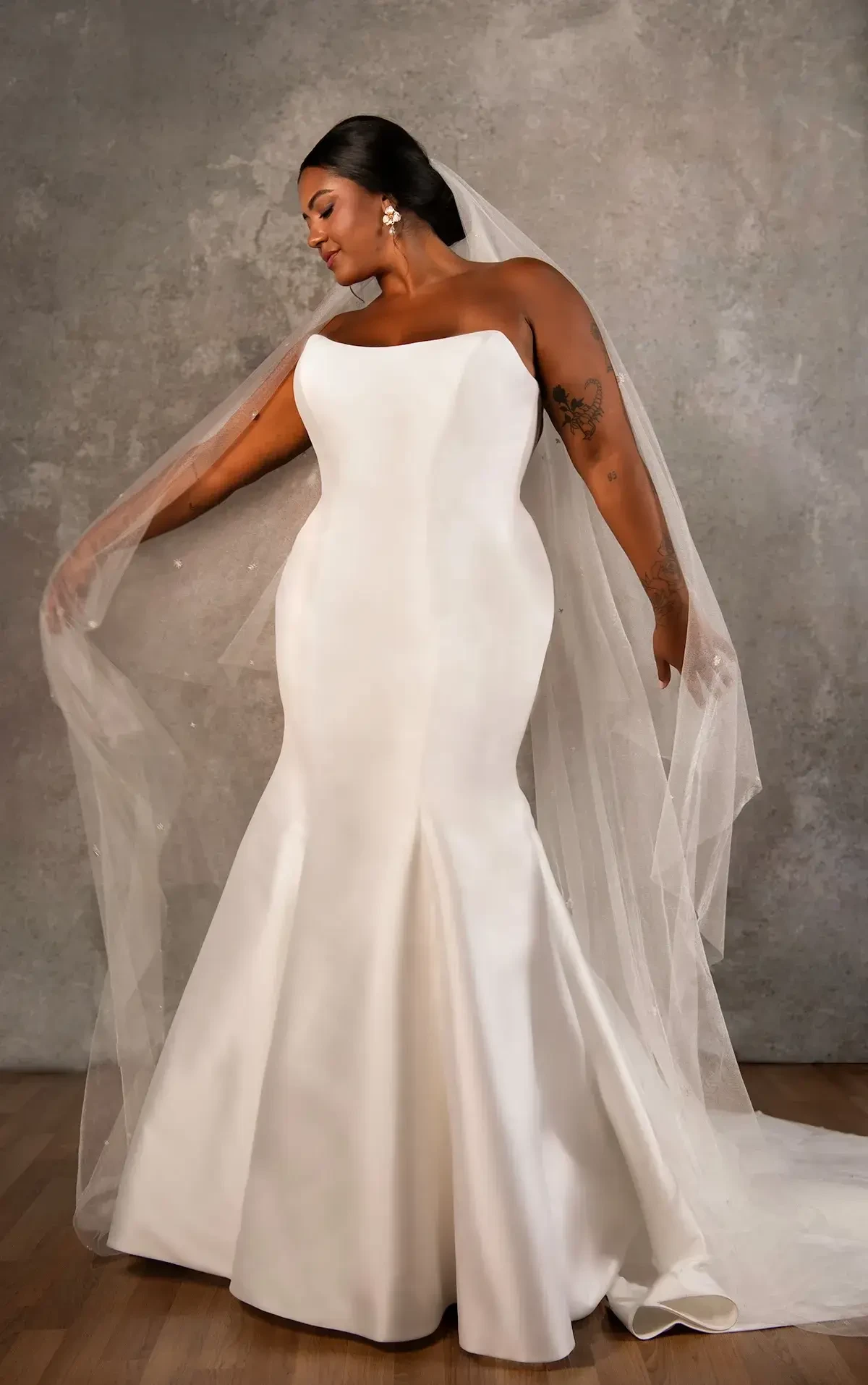 Bride in an elegant, strapless white wedding gown with a mermaid cut and sheer veil, posing gracefully against a textured gray backdrop.