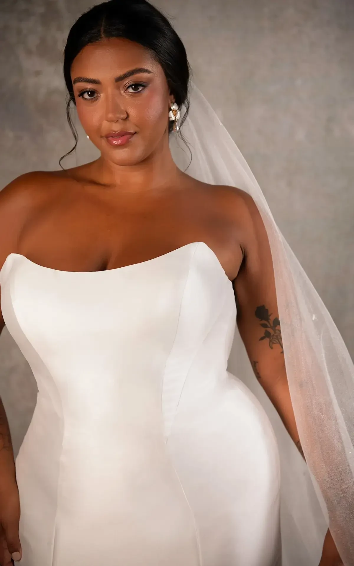 A bride in a strapless white wedding dress with a sheer veil smiles softly. She has floral tattoos on her arm. The background is a textured gray wall.