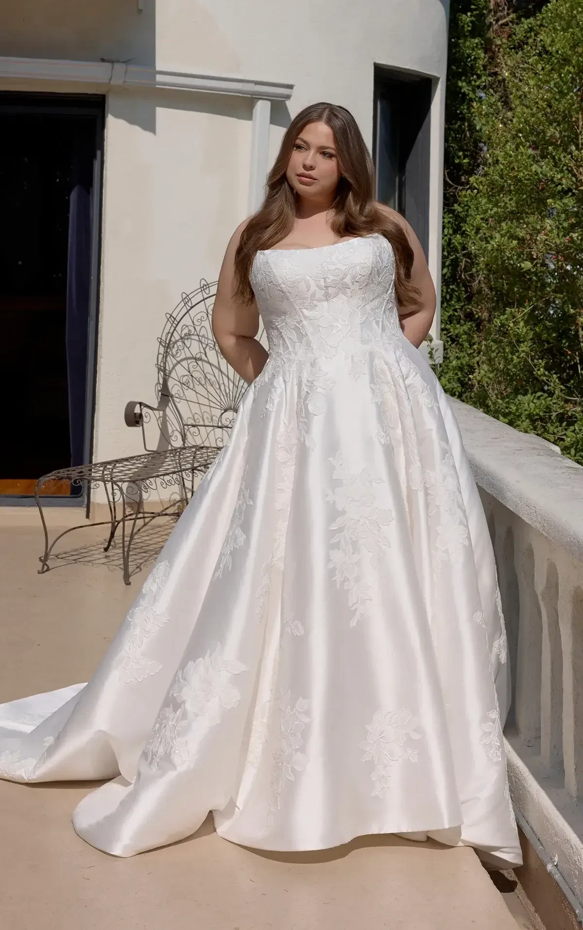 A woman in a white, elegant wedding gown stands on a balcony, surrounded by greenery.