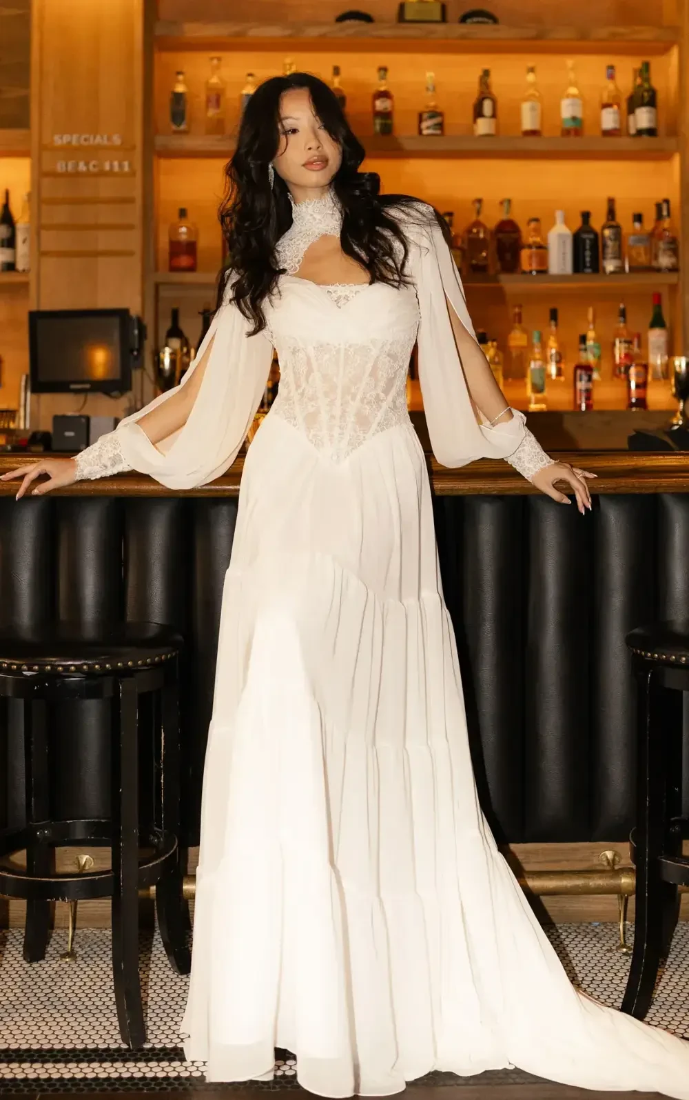 A woman wearing a white dress with long sleeves and a corset-style bodice poses in front of a bar with various bottles displayed in the background.