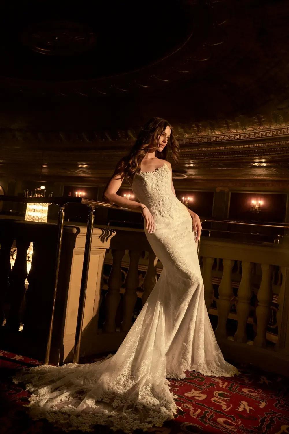 A model wearing a stunning lace wedding gown poses gracefully on a balcony in a dimly lit venue.