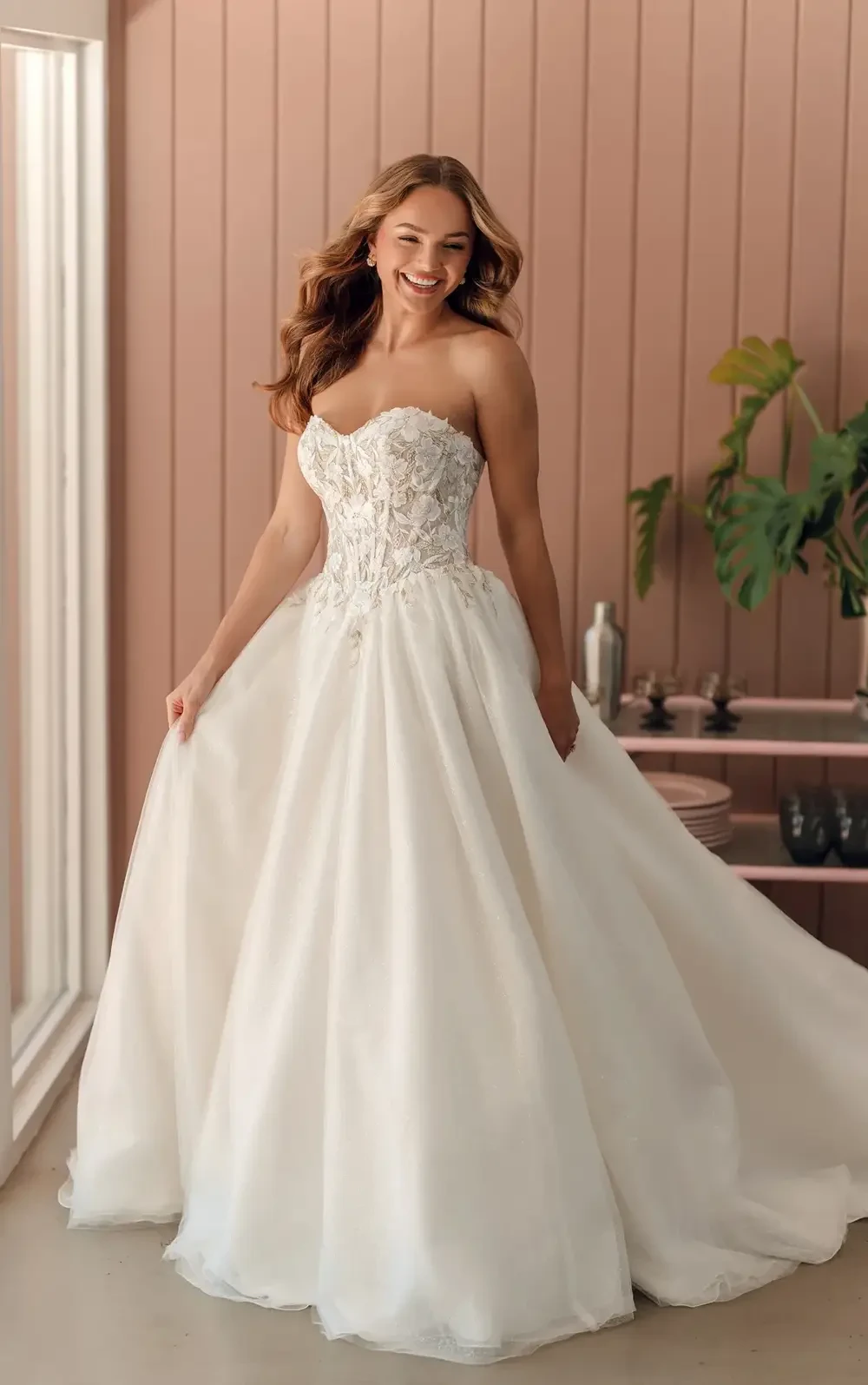 A smiling woman in a strapless white wedding dress, standing in a bright room with a pink wall and greenery.