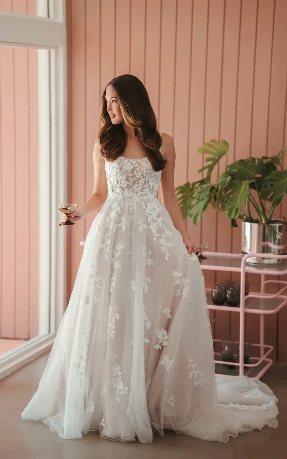 A woman in a floral-embellished, strapless wedding dress stands by a large window, holding a drink, with a pink wall and potted plants in the background.